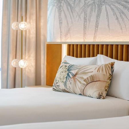 Amora Hotel Brisbane Bagian luar foto The photo shows a stylish hotel room featuring two neatly made beds with white linens. Each bed has several pillows, one of which has a decorative cover with a tropical plant pattern. Behind the beds is a modern headboard, and the wall is decorated w