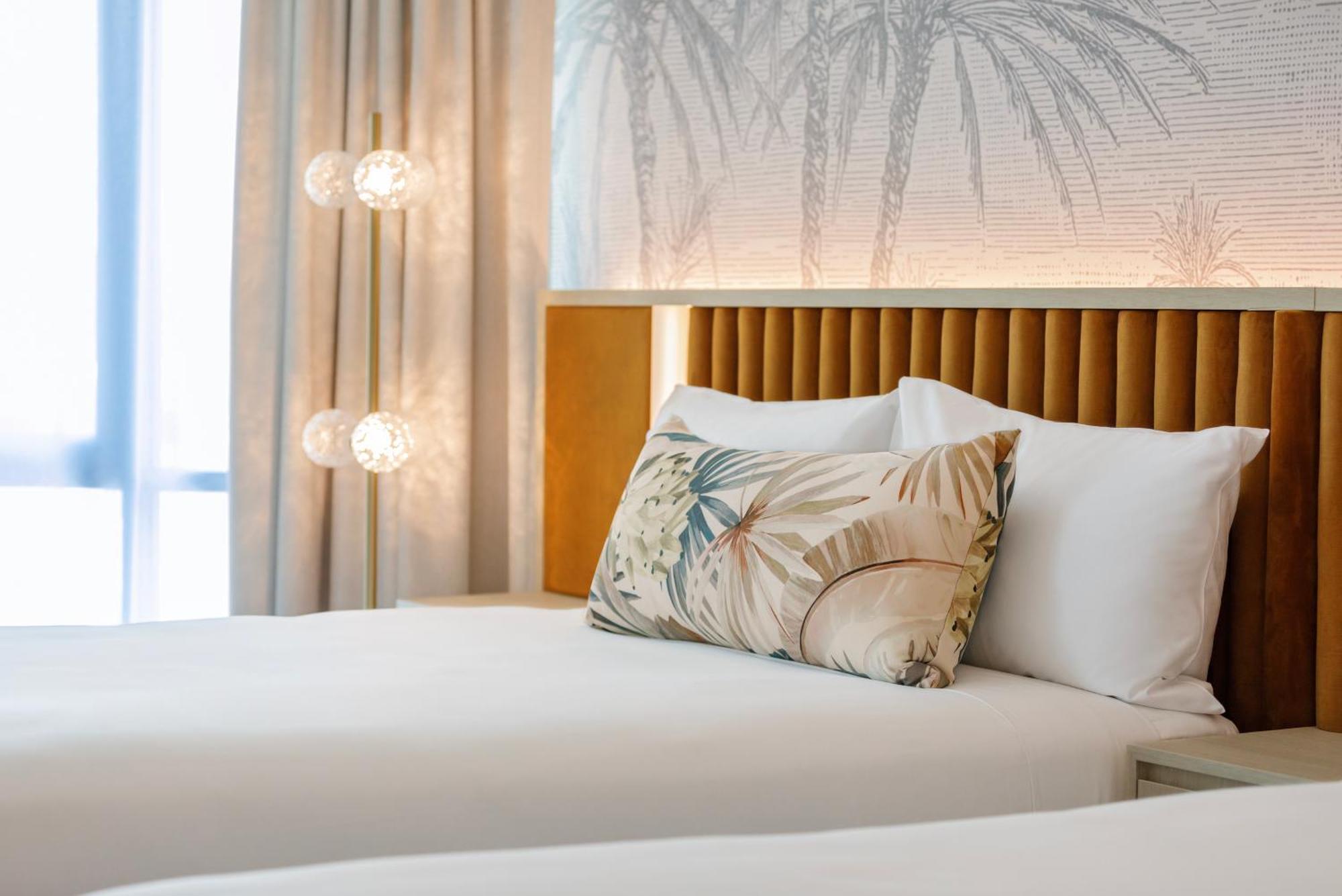 Amora Hotel Brisbane Bagian luar foto The photo shows a stylish hotel room featuring two neatly made beds with white linens. Each bed has several pillows, one of which has a decorative cover with a tropical plant pattern. Behind the beds is a modern headboard, and the wall is decorated w