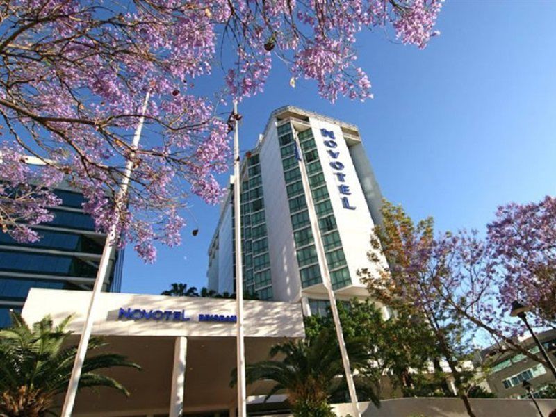 Amora Hotel Brisbane Bagian luar foto The photo shows a modern hotel building, identified as "Novotel," rising above a landscaped area with palm trees. In the foreground, there are vibrant purple flowering trees, likely jacarandas, which add to the picturesque scene. The sky is clear and